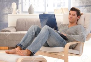 8398164-Goodlooking-young-man-relaxing-at-home-in-armchair-sitting-in-living-room-with-laptop-computer-smili-Stock-Photo